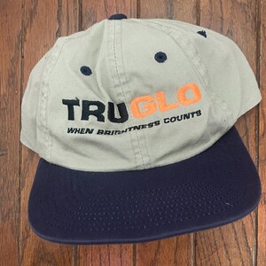 May include: A light gray baseball cap with a navy blue brim. The cap features the text "TRU GLO" in black and orange, with the tagline "WHEN BRIGHTNESS COUNTS" below. The cap has a curved brim and a button on top.