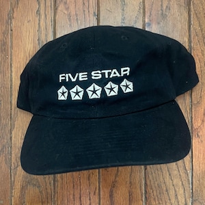 Puede incluir: Gorra de béisbol negra con el texto "FIVE STAR" en blanco sobre cinco emblemas de estrellas blancas. La gorra está hecha de un material suave y tiene una visera curva.