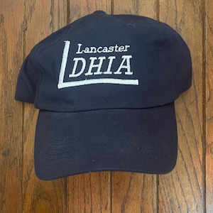 May include: Navy blue baseball cap with the text "Lancaster DHIA" embroidered in white. The cap has a curved brim and a structured crown. The hat is made of a durable fabric.