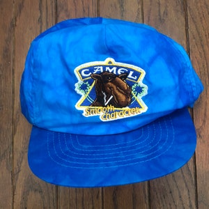 Könnte beinhalten: Eine leuchtend blaue Baseballkappe mit Batikmuster und einem zentralen gestickten Aufnäher. Der Aufnäher zeigt eine braune Kamelillustration, den Text CAMEL und die Worte Smooth character in Gelb auf einem blau-gelben Dreieck.