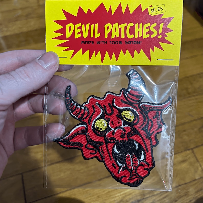 Op de afbeelding: Rode geborduurde strijkpatch met een duivelsgezicht met gele ogen. De patch is verpakt in een doorzichtige plastic zak met een gele en rode kop die "Devil Patches! Made with 100% Satan!" zegt en een prijskaartje van &euro; 6,66.