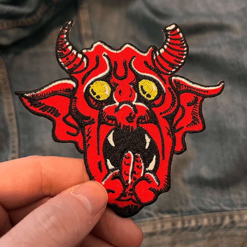 Op de afbeelding: Rode en zwarte geborduurde strijk-op patch van een cartoon duivel met gele ogen en een uitstekende tong.