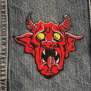 May include: Red devil with yellow eyes and a black mouth, giving the middle finger. The devil is on a blue denim background.