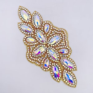 May include: A gold and iridescent crystal embellishment with a floral design. The embellishment is made of small, round crystals and larger, teardrop-shaped crystals.