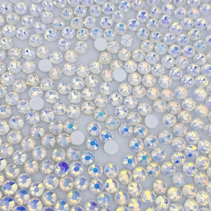 May include: A close-up image of a large quantity of iridescent white rhinestones. The rhinestones are scattered on a white surface.