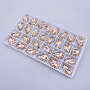 May include: A white tray filled with 24 clear, faceted, oval-shaped rhinestones. The rhinestones have a light champagne color.