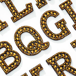 Pu&ograve; includere: Un set di lettere dorate in strass, tra cui le lettere B, G, I, L, Q e R. Le lettere sono disposte in modo casuale su uno sfondo bianco.