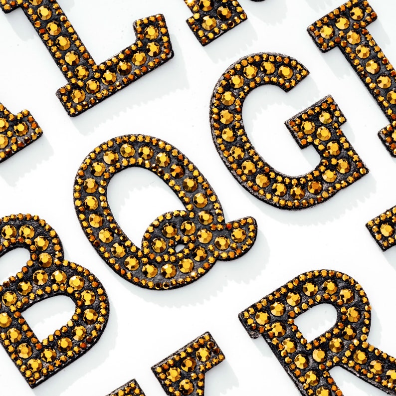 Pu&ograve; includere: Un set di lettere dorate in strass, tra cui le lettere B, G, I, L, Q e R. Le lettere sono disposte in modo casuale su uno sfondo bianco.