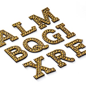 Pu&ograve; includere: Un set di lettere in strass dorate, tra cui A, L, M, B, Q, G, I, X, R ed E. Le lettere sono realizzate in tessuto nero con strass dorati.