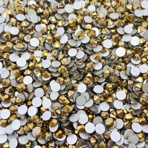 May include: A large quantity of gold-colored rhinestones with flat backs. The rhinestones are scattered on a white surface.