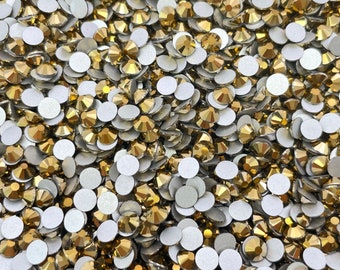 May include: A large quantity of gold-colored rhinestones with flat backs. The rhinestones are scattered on a white surface.
