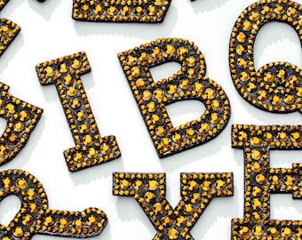 Peut inclure: Un ensemble de lettres en strass dorées, comprenant les lettres B, E, I, L, Q, R et X. Les lettres sont sur un fond blanc.