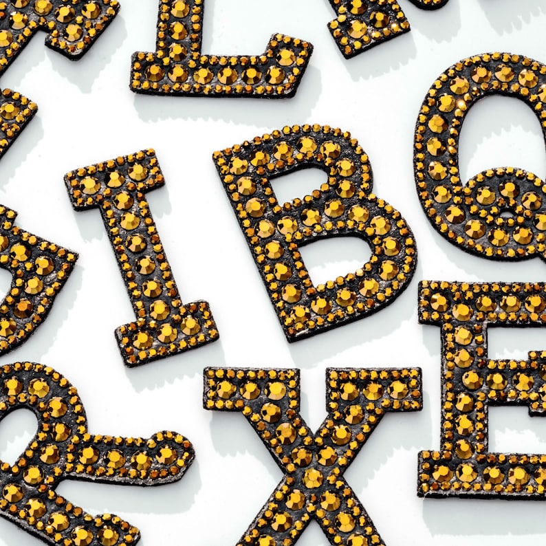 Pu&ograve; includere: Un set di lettere in strass dorate, tra cui le lettere B, E, I, L, Q, R e X. Le lettere sono su uno sfondo bianco.