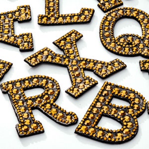 Pu&ograve; includere: Un set di lettere dorate in strass, tra cui le lettere B, L, Q, R e X. Le lettere sono su uno sfondo bianco.