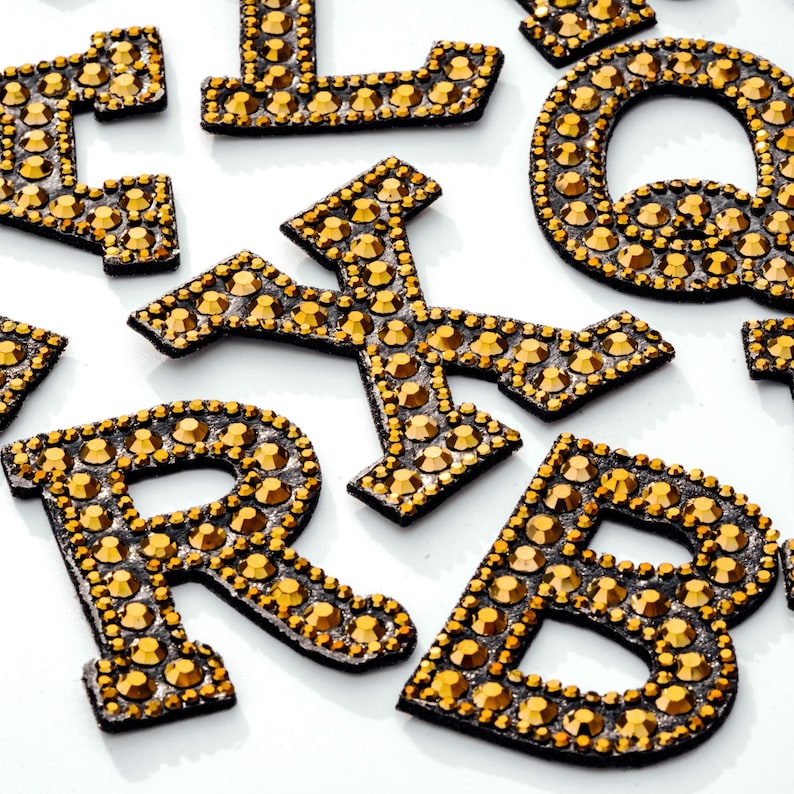 Pu&ograve; includere: Un set di lettere dorate in strass, tra cui le lettere B, L, Q, R e X. Le lettere sono su uno sfondo bianco.