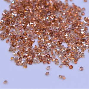 May include: A scattering of small, faceted gemstones in shades of amber and light brown. The gems are scattered across a white surface, catching the light and creating a sparkling effect. The gems are likely used for jewelry making or other decorative purposes.