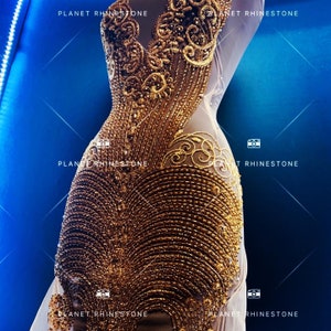May include: A white dress with gold embellishments. The dress is covered in small gold beads and sequins, creating a shimmering effect. The dress has a fitted bodice and a flowing skirt.