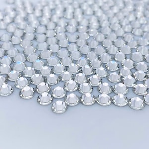 May include: A close-up of a large quantity of clear, faceted rhinestones. The rhinestones are arranged in a random pattern and are scattered across a white surface.