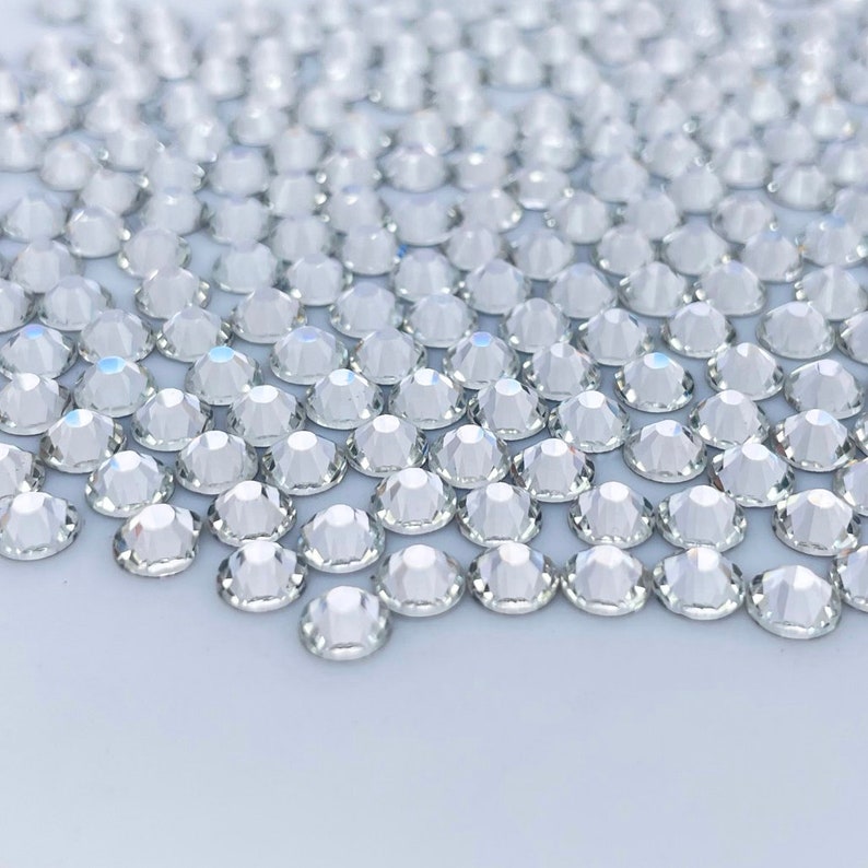 May include: A close-up of a large quantity of clear, faceted rhinestones. The rhinestones are arranged in a random pattern and are scattered across a white surface.