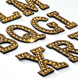 Pu&ograve; includere: Lettere dorate dell'alfabeto, fatte di strass, su uno sfondo bianco. Le lettere sono disposte in ordine casuale, con alcune sovrapposte.