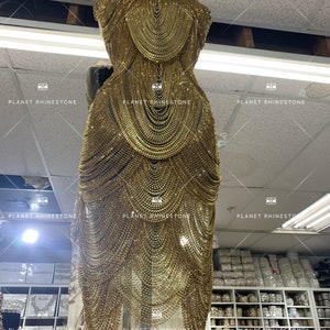 May include: A gold-colored beaded dress with a chain-like design. The dress is on a mannequin and is displayed in a store with shelves of beads and rhinestones.