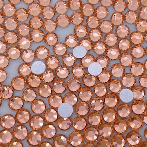 May include: A close-up image of a large quantity of small, round, peach-colored rhinestones. The rhinestones are arranged in a random pattern and are scattered across a white surface.