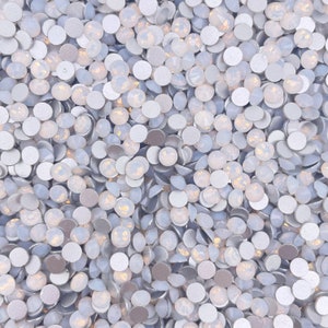 May include: A close-up view of a large quantity of small, round, iridescent white rhinestones. The rhinestones have a silver backing and are scattered randomly.