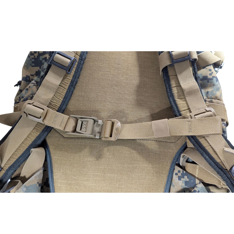 Op de afbeelding: Een camouflage rugzak in tan en groen met een zwarte gesp en een bruine band met een gesp. De band is verstelbaar en heeft een zwarte gesp.