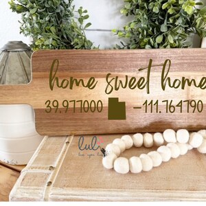 May include: Wooden chopping board with a handle, featuring the text "home sweet home" and coordinates "39.977000 -111.764790". The board is brown and has a natural wood grain.