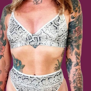 May include: A two-piece lingerie set featuring a snakeskin pattern in shades of gray and white. The set includes a triangle-style bra and high-waisted bottoms, both trimmed with white edging. The background is a solid dark purple.