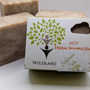 May include: A bar of brown soap with a white label that says "WILDLAND" and "ACV HERBAL SHAMPOO BAR" with a green tree and a person silhouette. A smaller white label with green text says "Ecklonia Cava".