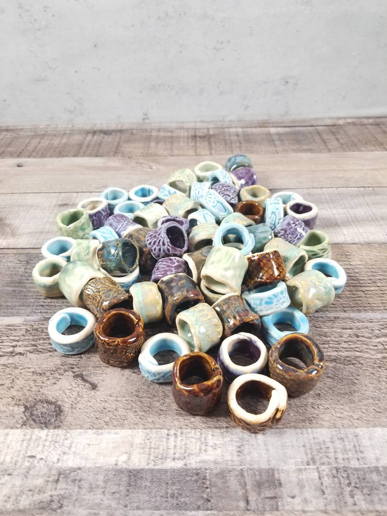 Pu&ograve; includere: Un mucchio di anelli in ceramica di vari colori, tra cui blu, verde, marrone e viola. Gli anelli sono di diverse dimensioni e forme e sono disposti in un motivo casuale.