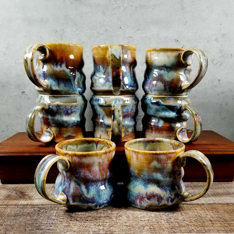 May include: Set of eight ceramic mugs with a unique, swirled glaze in shades of blue, brown, and gold. The mugs have a textured, uneven surface and are stacked in pairs.