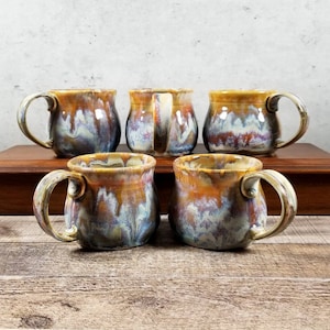 May include: Five ceramic mugs with a brown, white, and blue glaze. The mugs have a rounded body and a handle.