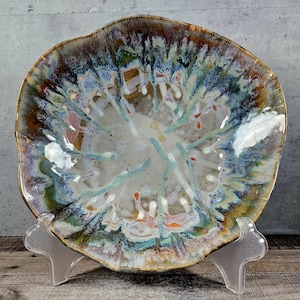 May include: A large, ceramic bowl with an abstract, colorful glaze. The bowl has a unique, organic shape and is decorated with shades of blue, green, brown, and white.
