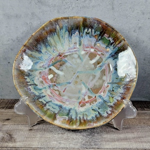 May include: A large, ceramic serving bowl with a unique, abstract design. The bowl has a white base with streaks of blue, green, and pink. The rim of the bowl is a golden brown color.