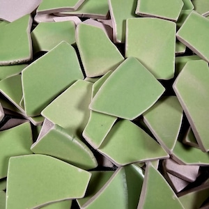 May include: Close-up of a pile of light green ceramic tiles with irregular shapes. The tiles have a glossy finish and are scattered, revealing a light-colored backing. The edges of the tiles are slightly rounded, and the overall effect is a mix of geometric and organic forms.
