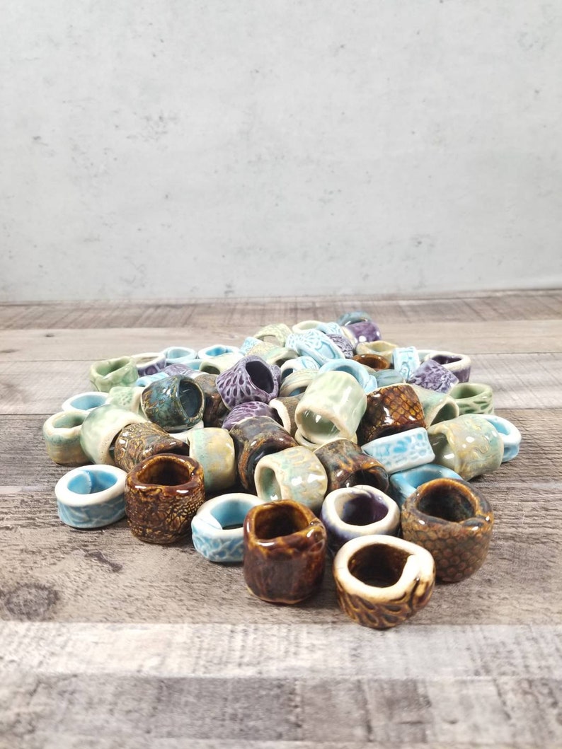 Pu&ograve; includere: Un mucchio di anelli in ceramica colorati, alcuni con una superficie testurizzata, in tonalit&agrave; di verde, blu, marrone e viola.