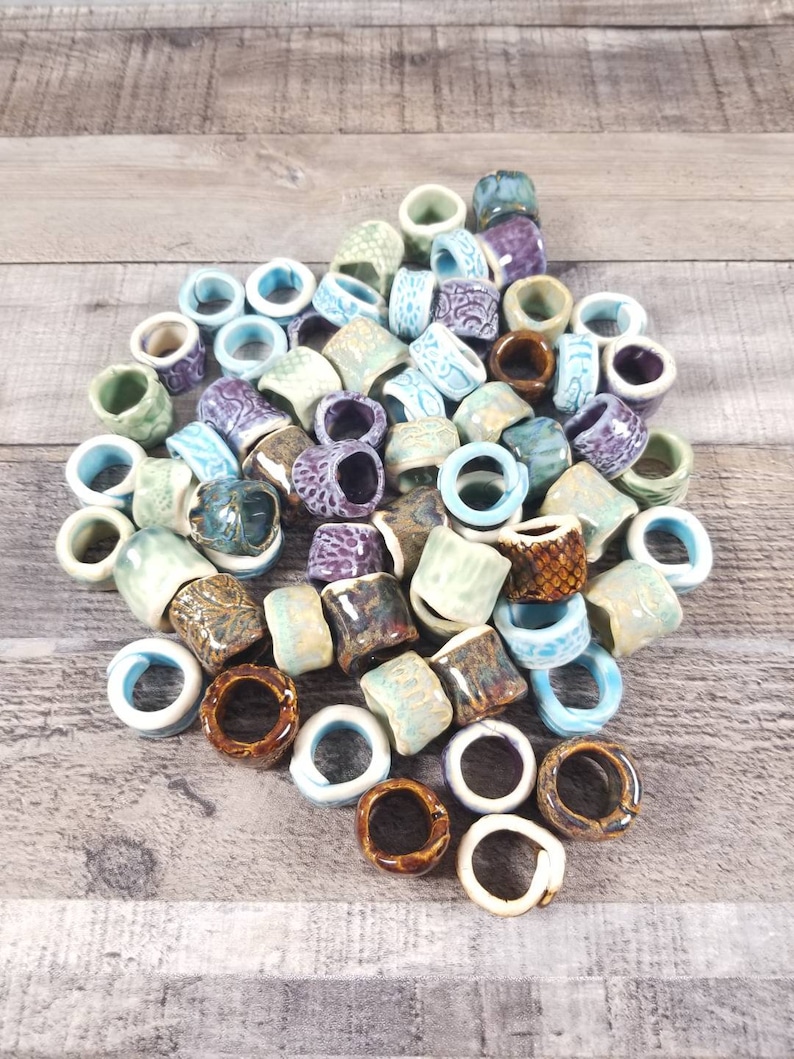 Pu&ograve; includere: Una collezione di anelli in ceramica di vari colori e motivi, tra cui blu, verde, marrone e viola. Gli anelli sono impilati e sparsi su una superficie di legno.
