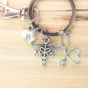 May include: Silver keychain with a heart charm with the letter "H", a medical symbol charm with "MDA" text, and a stethoscope charm. The keychain has a lobster claw clasp and a large ring.