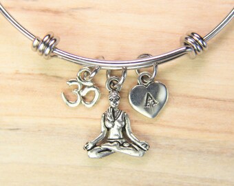 May include: A silver bangle bracelet with three charms: a yoga pose charm, an Om symbol charm, and a heart charm with the letter A.