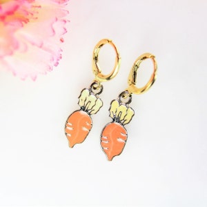 May include: A pair of gold hoop earrings with a dangling carrot charm. The carrot is orange with a yellow top and green leaves.