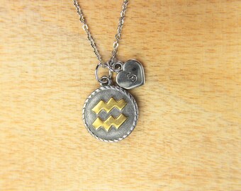May include: Silver chain necklace with a silver heart charm with the letter 'S' and a round silver charm with a gold Aquarius zodiac symbol.