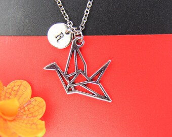 May include: Silver necklace with a silver origami crane pendant and a small round charm with the letter "R" on it. The necklace is on a red background.