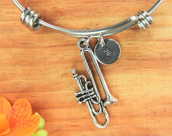 May include: A silver bangle bracelet with a charm of a trombone and a silver charm with the letter K. The bracelet is on a wooden surface.