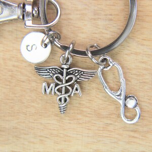 May include: Silver keychain with a medical caduceus charm, a stethoscope charm, and a small round charm with the letter 'S' on it.