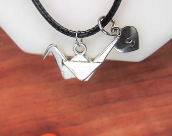 May include: A silver origami crane charm necklace with a small silver heart charm with the letter 'S' on it. The necklace is on a black cord.