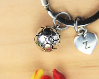 May include: A silver-toned bracelet with a soccer ball charm and a heart-shaped charm with the letter "Z". The bracelet has a black band and is set against a light brown wooden surface. The soccer ball charm is detailed.