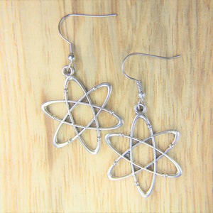 May include: A pair of silver-colored earrings shaped like atom models. Each earring features a hook for pierced ears and a star-like design with intersecting lines, set against a light brown wooden surface.