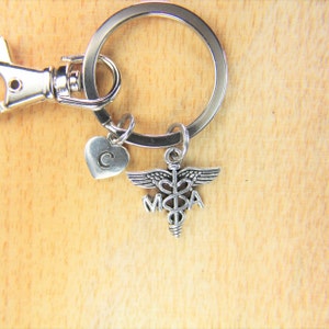 May include: Silver keychain with a medical symbol charm and a heart charm with the letter C. The keychain is attached to a silver key ring.